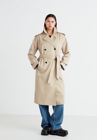 Abrigo trench de doble botonadura en beige con cintura ceñida por un cinturón, amplios solapas y botones negros. El modelo lleva pantalones de pierna ancha y zuecos.