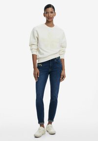 Sweatshirt bianco con design floreale testurizzato, abbinato a jeans blu scuro con ricami colorati in vita e scarpe bianche.