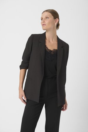 Černý blazer s úzkým střihem, má jedno tlačítko, klopové límce a ohrnuté rukávy, spárovaný s černým krajkovým topem.