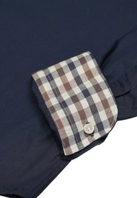 Camicia navy con polsini a scacchi nei toni crema, marrone e grigio, con un singolo bottone decorativo e texture morbida.