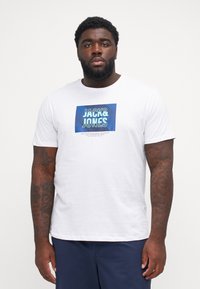 Λευκό κοντομάνικο μπ�λουζάκι με μπλε γραφικό ορθογώνιο που περιέχει το κείμενο "JACK & JONES", με φόντο υφής.
