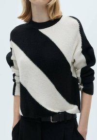 Pull en tricot noir et crème avec des rayures diagonales, encolure ronde et poignets côtelés, associé à une ceinture noire sur un pantalon sombre.