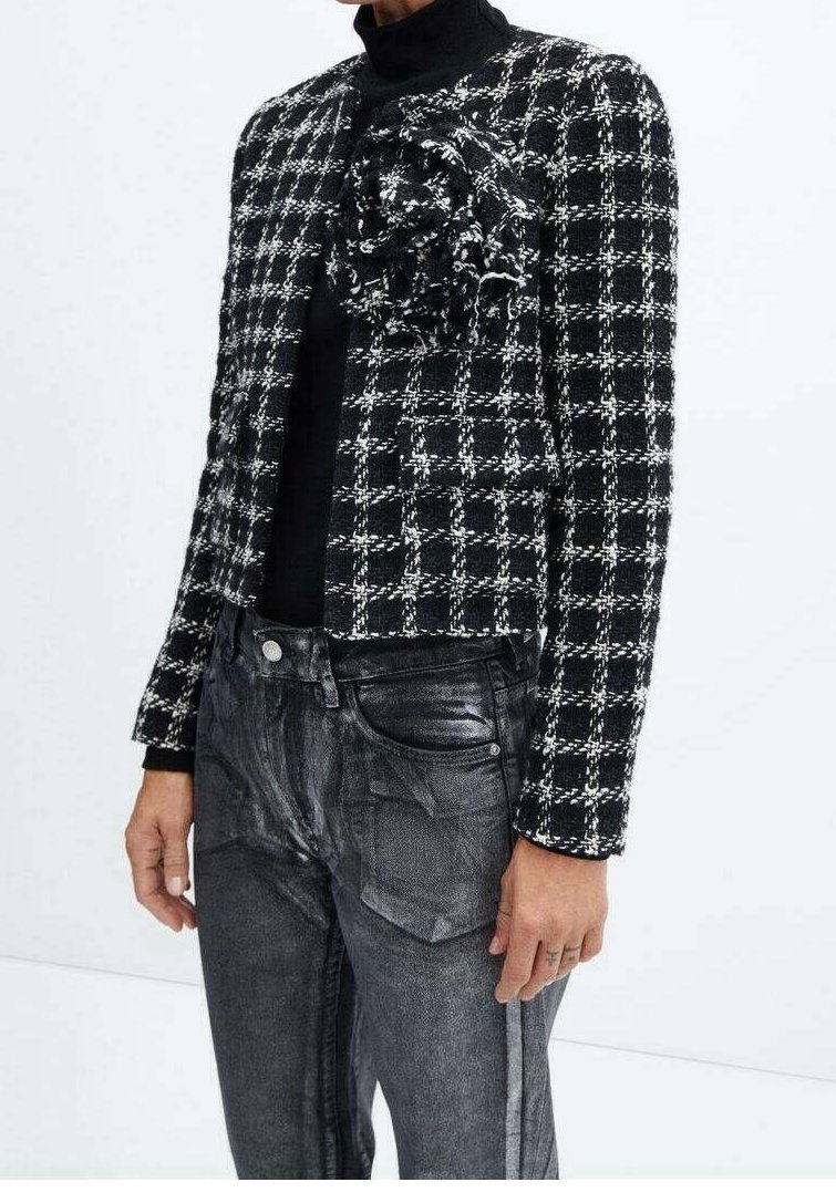 Sort og hvid tweedjakke med en tætsiddende pasform, en struktureret blomsteraccent på venstre side, parret med mørke, slidte jeans.