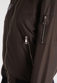 Giacca bomber marrone con una texture liscia, dotata di una tasca con zip sulla manica e dettagli in metallo argentato.