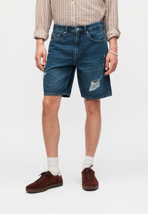 Μπλε denim σορτς μέσης μήκους με φθαρμένες λεπτομέρειες, συνδυασμένα με πουκάμισο καρώ, λευκές κάλτσες και καφέ αθλητικά παπούτσια.