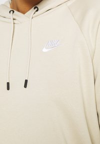 Khaki hoodie i mjukt tyg; har en dragsko i huva och en vit broderad Nike-logotyp på bröstet.