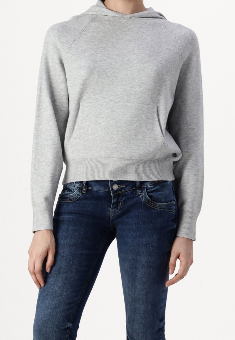 Femme portant un sweat à capuche gris clair avec poche avant et un jean moulant bleu foncé, se tenant devant un fond uni.