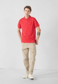 Rood poloshirt met korte mouwen en een kraag, gecombineerd met beige cargobroek met zakken en opgerolde pijpen, gedragen met witte sneakers.