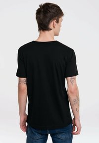 Black cotton t-shirt with short sleeves, round neckline, and a smooth texture, shown from the back. Visible tattoos on left forearm.