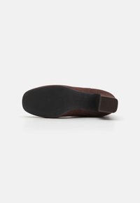 Zapato de ante marrón con puntera redonda y suela de goma negra texturizada. Presenta un tacón bajo y un detalle de costura mínima.