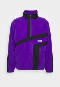 Lilla fleece-pullover med en sort lynlås, der har et halvlynlåsdesign, sorte accenter og et broderet logo på forsiden.