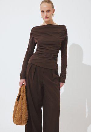 Femme portant un haut marron à manches longues froncées et un pantalon marron à jambes larges, tenant un sac à main en paille beige, debout devant un fond blanc uni.