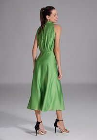 Grünes Satin-Midikleid mit hohem Kragen und geraffter Taille. Es hat einen fließenden Rock und wird mit schwarzen und goldenen High-Heels kombiniert.