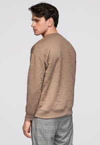 Nuori mies sivuprofiilissa, päällään teksturoitu ruskea collegepaita kohokuvioiduilla kirjaimilla ja harmaat ruudulliset housut, taustalla yksivärinen tausta.