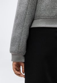 Main d'une personne reposant le long du corps, portant un pull gris à manches longues avec un poignet côtelé et un pantalon ou une jupe noire.