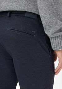 Primo piano di una persona che indossa pantaloni scuri a trama e un maglione grigio a maglia, che mostra la tasca posteriore e parte della mano destra.