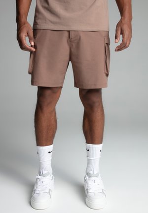Mand iført brune cargoshorts, hvide Nike-strømper og hvide sneakers, står mod en neutral baggrund.