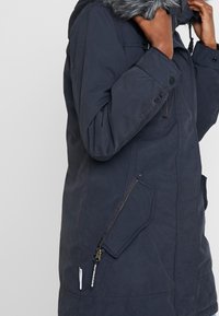 Personne portant un manteau d’hiver bleu foncé avec une capuche bordée de fourrure, tenant la capuche près de son visage.