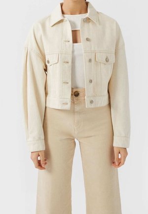 Persoon draagt een lichtbeige cropped spijkerjack over een wit geribd topje met een uitgesneden detail, gecombineerd met beige broeken met hoge taille.