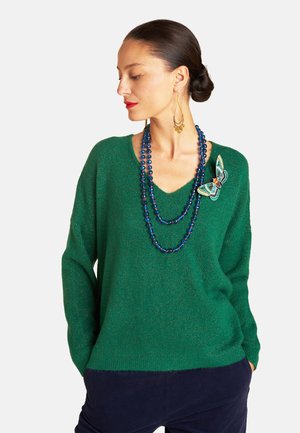 Grüner, glitzernder Pullover mit V-Ausschnitt, der ein besticktes Schmetterlingsdetail auf der linken Seite aufweist. Passt zu geschichteten blauen Perlenhalsketten.