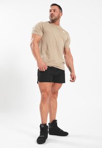 Šviesiai rudos spalvos trumpomis rankovėmis marškinėliai, juodos sportinės šortai ir juodi aukšti sportbačiai. Raumeninga figūra, stovima poza su atsipalaidavusiomis rankomis. Balto logotipo detalė.