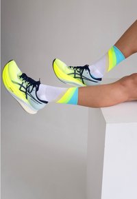Bright yellow athletic shoes with mesh upper and black accents, paired with colorful patterned socks in blue and green, worn on a white surface.