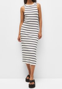 Femme portant une robe midi blanche sans manches avec des rayures horizontales noires et des sandales compensées noires, se tenant devant un fond blanc uni.