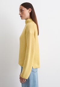 Gelber Strickpullover mit hohem Kragen und langen Ärmeln. Hat ein strukturiertes Design und seitliche Schlitze. Getragen mit hellblauen Jeans.