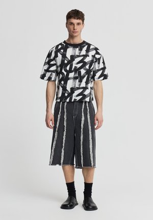 Modello maschile in piedi indossa una t-shirt oversize con stampa astratta in bianco e nero, pantaloncini neri al ginocchio con strisce verticali di frange, calzini neri e scarpe nere.
