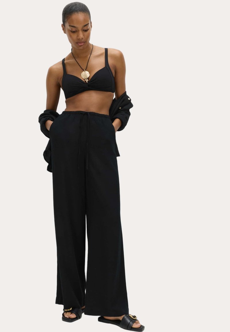 Mujer vestida con pantalones anchos negros, top corto negro, camisa negra abierta, collar con colgante y sandalias negras, de pie con las manos en los bolsillos.