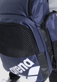 Mørkeblå rygsæk med struktureret sort mesh-panel, forstærkede lynlåse og fremtrædende hvidt logo. Lavet af slidstærkt stof med flere lommer.