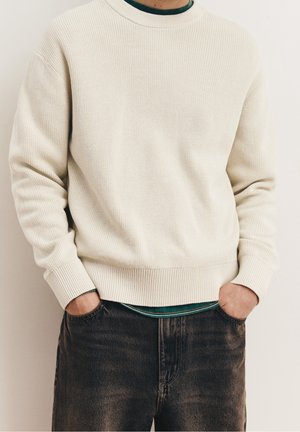 Persona que lleva un suéter de punto crema sobre una camisa verde oscuro, con las manos en los bolsillos de unos vaqueros negros desgastados.