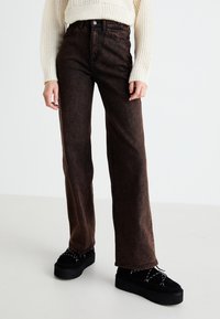 Jeans de perna larga castanhos, feitos de denim, com um corte alto, leves desbotados e bolsos frontais, combinados com sapatos plataforma pretos.