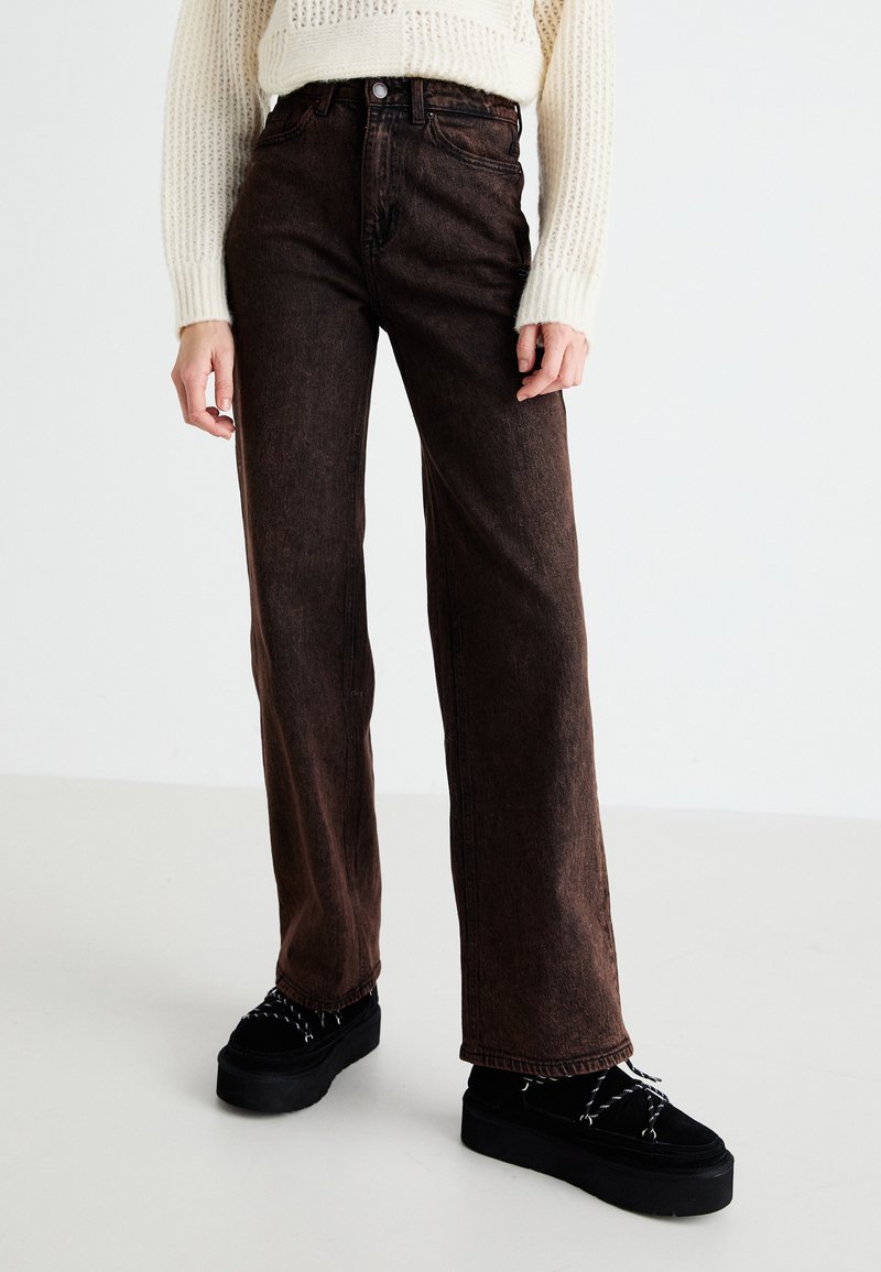 Jeans de perna larga castanhos, feitos de denim, com um corte alto, leves desbotados e bolsos frontais, combinados com sapatos plataforma pretos.