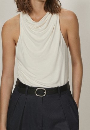 Kobieta w białej, bezrękawnikowej bluzce z luźnym kołnierzem, wsuniętej w ciemne, wysokie spodnie z czarnym paskiem i srebrną sprzączką.