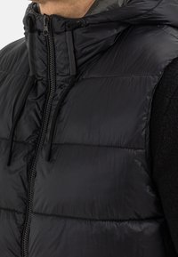 Schwarze Pufferweste mit einem Reißverschluss vorne, verstellbarem Kapuzenaufsatz und strukturiertem Stoff. Merkmale sind horizontale Nähte und glattes Futter.