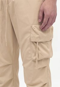 Main d’homme reposant près de la poche d’un pantalon cargo beige avec rabat et détail cordon sur fond blanc.