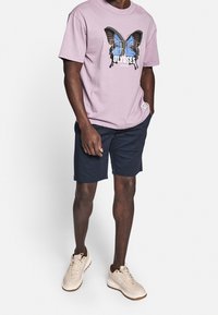 T-shirt violet clair avec un graphique de papillon, short bleu marine et baskets beiges. Le T-shirt a un col rond et une coupe décontractée.