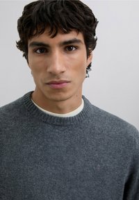 Suéter gris claro con cuello redondo acanalado, textura suave y un sutil detalle con el logo en el pecho, llevado sobre una camisa blanca.