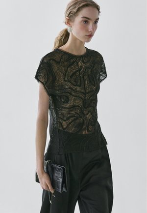 Jonge vrouw met licht haar in een staart, draagt grote zilveren oorbellen, een zwarte doorzichtige top met textuur, zwarte broek en houdt een zwarte clutch vast.