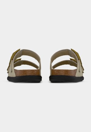 Par beige sandaler med to remme og guldfarvede spænder, korkindlæg og sorte såler, vist bagfra mod hvid baggrund.