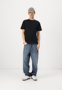 Svart bomullst-skjorte kombinert med lyseblå denimjeans, hvite og grå sneakers og en grå caps, stående mot en ensfarget bakgrunn.