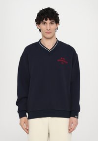 Sweatshirt azul-marinho com decote em V, apresentando acabamentos brancos, bordado vermelho "MILANO GYMNASIUM EA7" e punhos e bainha canelados.