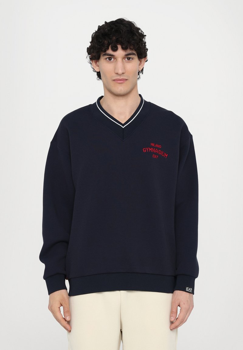 Sweatshirt azul-marinho com decote em V, apresentando acabamentos brancos, bordado vermelho "MILANO GYMNASIUM EA7" e punhos e bainha canelados.