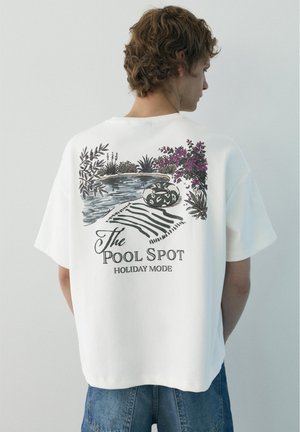 Jonge man met wit T-shirt met illustratie van zwembad, planten, opblaasbaar, handdoek en tekst "The Pool Spot Holiday Mode" op de rug