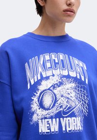 Personne portant un sweat bleu avec un texte blanc "NIKECOURT NEW YORK" et un dessin d'une balle de tennis frappant un filet avec un logo Nike.