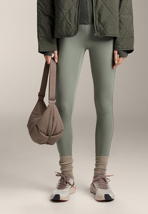 Személy zsályazöld leggingset, steppelt sötétzöld dzsekit, bézs összegyűrt zoknikat és fehér tornacipőt visel, oldalán barna párnázott kézitáskát tart.