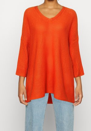 Femme portant un pull en laine ample orange vif à manches 3/4 et un jean bleu clair, debout devant un fond blanc.