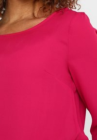 Blouse magenta avec un décolleté arrondi et des manches longues. Fabriquée en tissu léger et doux, présentant un drapé subtil et sans fermetures visibles.