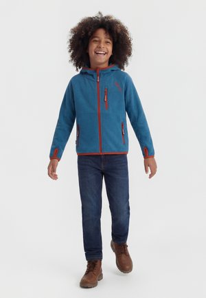 Smaidīgs bērns ar cirtainiem matiem, valkājot zilu džemperi ar rāvējslēdzēju, tumšas džinsas un brūnas zābaciņus, staigājot uz priekšu uz vienkārša fona.